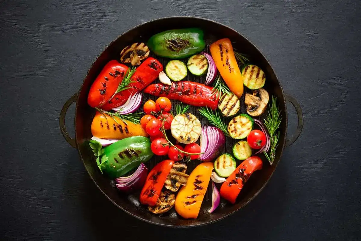 Un assortimento colorato di verdure grigliate, tra cui peperoni, zucchine, cipolle rosse, pomodorini e funghi, disposte in una padella di ghisa.