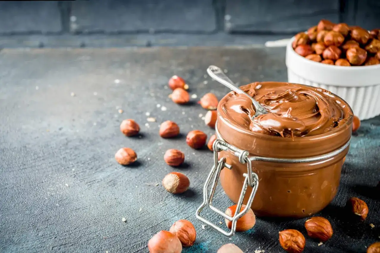 Un vasetto di crema di nocciole fatta in casa, con nocciole sparse intorno. Un cucchiaino affonda nella golosa crema.
