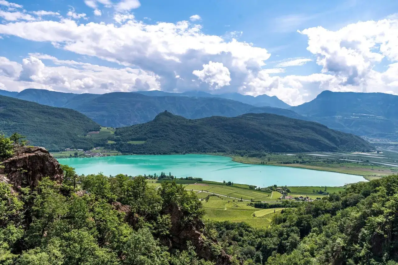 Panoramica mozzafiato del lago di Caldaro, con le sue acque turchesi che riflettono il cielo azzurro e le montagne verdi. Vigneti rigogliosi abbracciano la riva.