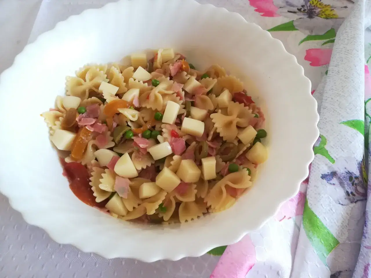 Insalata di pasta fredda con farfalle, cubetti di formaggio, prosciutto, piselli e pomodorini. Un'idea fresca e gustosa per un pranzo estivo.