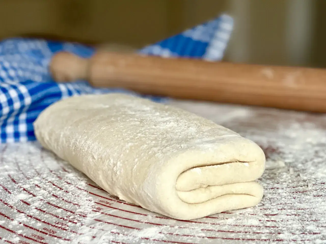 Impasto per ricetta pasta sfoglia, piegato e infarinato, pronto per essere steso. Un mattarello e un canovaccio a quadretti blu sullo sfondo.