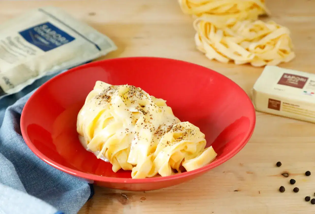 Un piatto di fettuccine alfredo cremose, condite con pepe nero macinato fresco, su un piatto rosso. Ingredienti per un'ottima pasta.