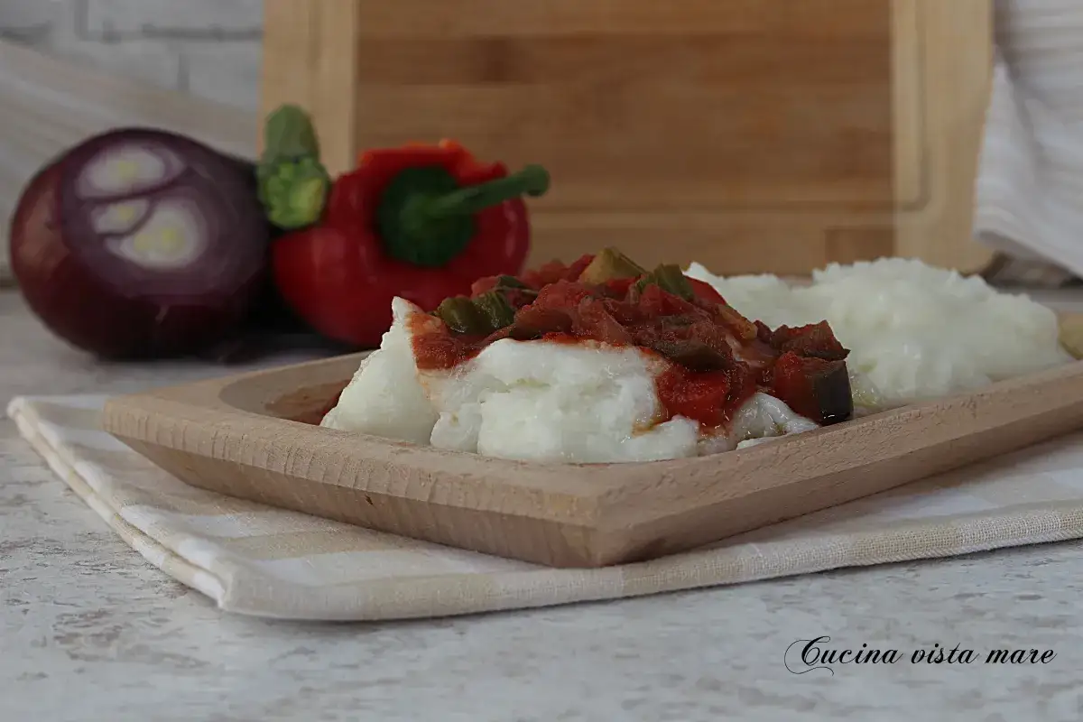 Polenta bianca servita in un piatto di legno con un contorno di verdure e pomodoro. Sullo sfondo, cipolla rossa e peperone.