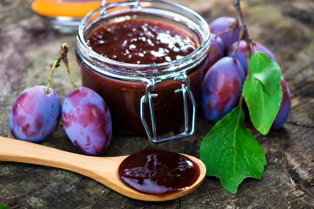 Un vasetto di marmellata di susine fatto in casa, con susine fresche e un cucchiaio pieno di questa delizia.