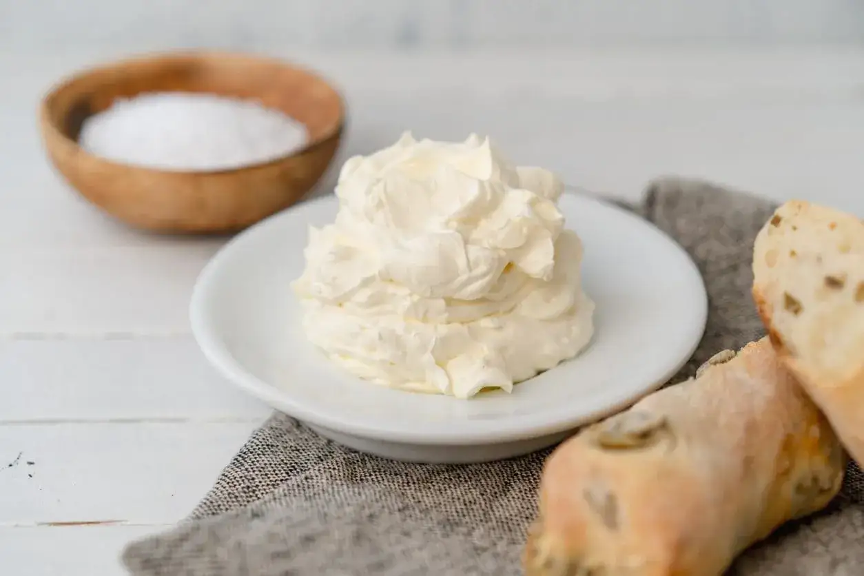 Un soffice cumulo di burro montato su un piattino bianco, pronto per essere spalmato su pane croccante. Accanto, una ciotola di sale grosso.