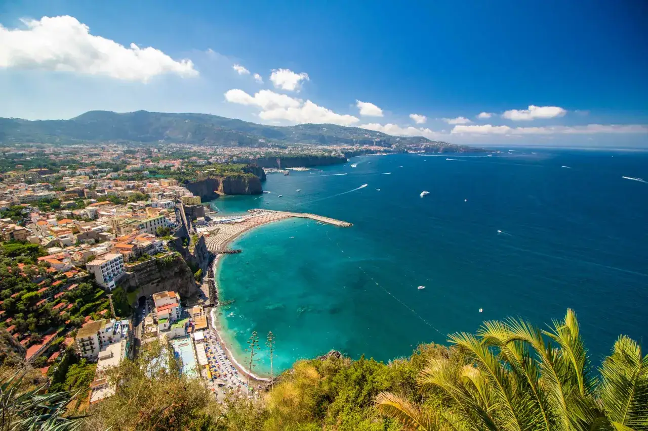 Vista panoramica della penisola sorrentina con case colorate a picco sul mare turchese, spiagge affollate e montagne sullo sfondo.