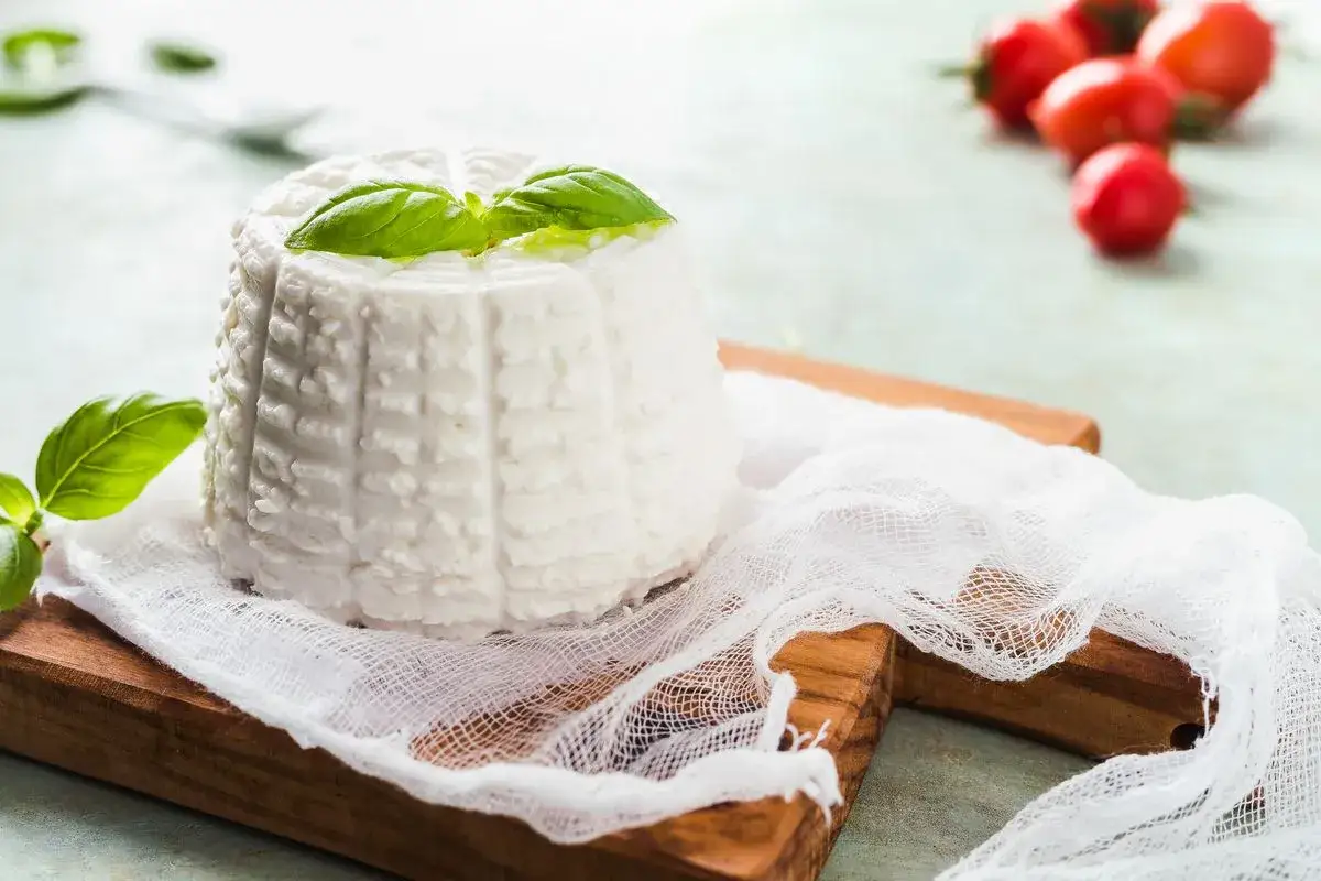 Ricotta fatta in casa, fresca e profumata, guarnita con basilico su un tagliere di legno. Pomodorini sfocati sullo sfondo.
