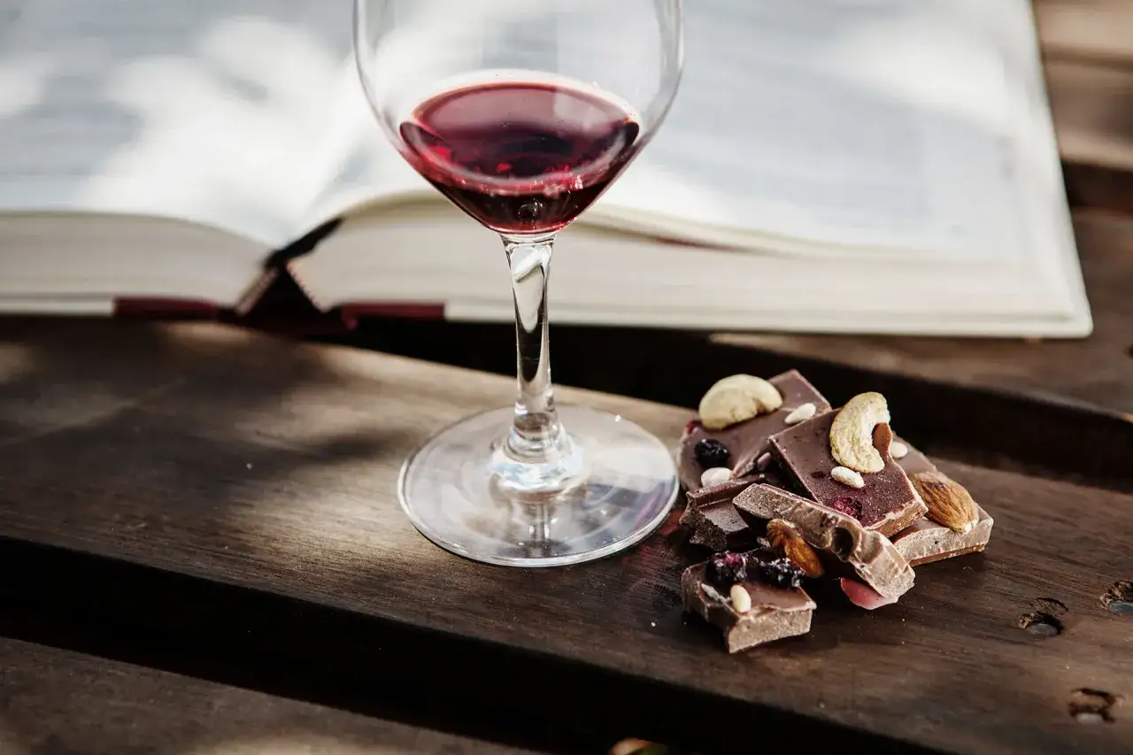Calice di vino rosso accanto a pezzi di cioccolato con frutta secca e noci. Un libro aperto sullo sfondo suggerisce un momento di relax con un perfetto abbinamento vino e cioccolato.